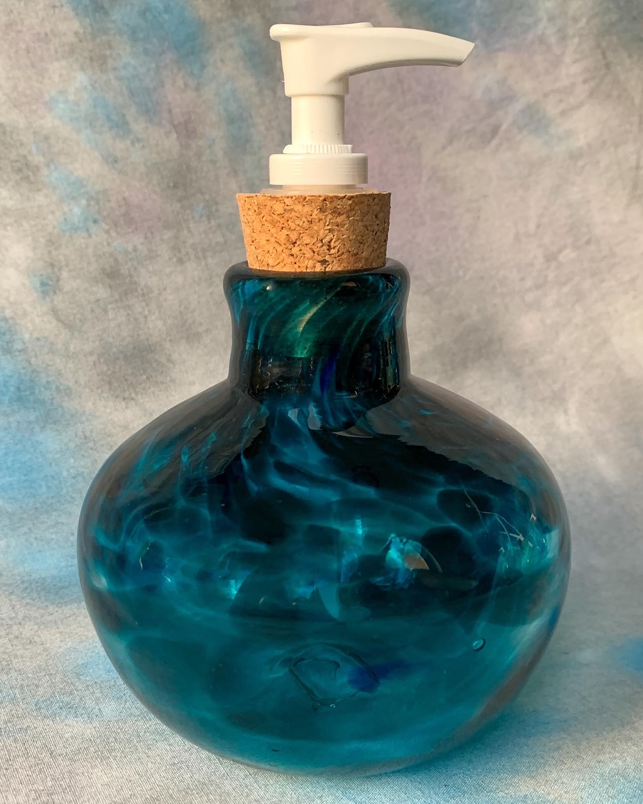 A round teal glass dispenser with swirling colour and a white pump